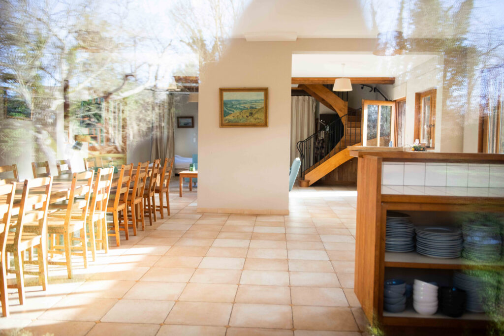 Intérieur lumineux du gîte, avec une grande table en bois, des chaises, et un escalier en bois menant à l'étage.