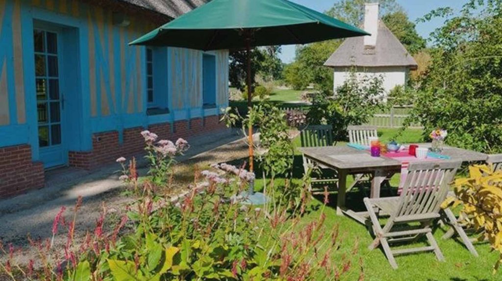 Terrasse ensoleillée avec mobilier en bois, parasol et fleurs, devant la chaumière Les Noyers en Normandie.