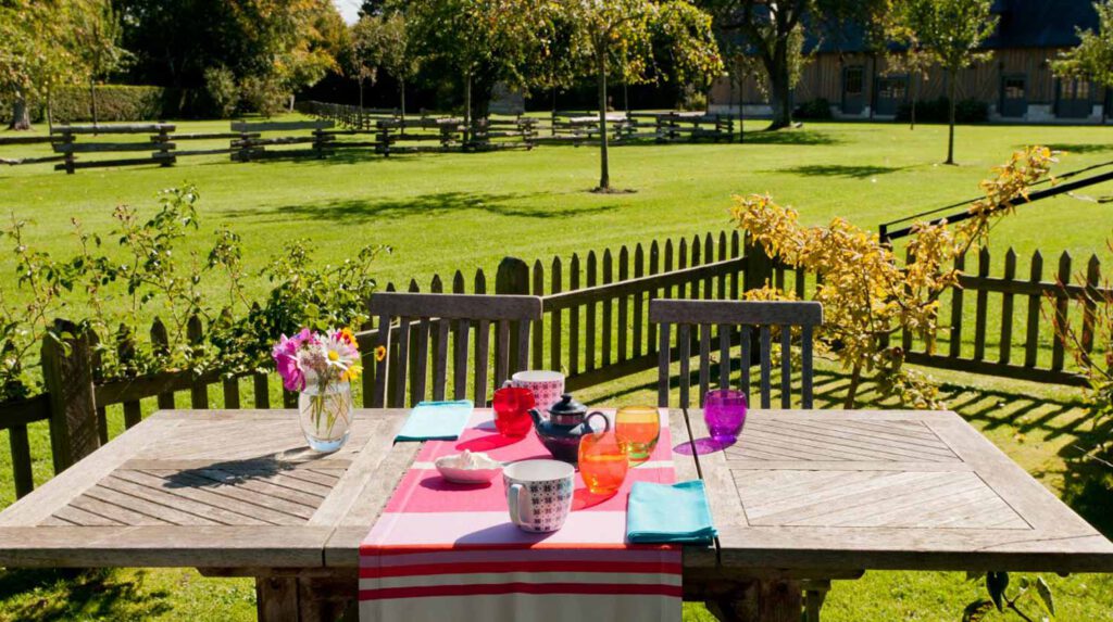 Table en bois avec nappes colorées, verres colorés et fleurs, dans un jardin verdoyant entouré de clôtures en bois.