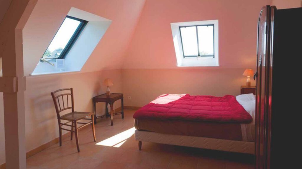 Chambre lumineuse avec lit à couverture rouge, chaise en bois, table de nuit et fenêtres à toit.